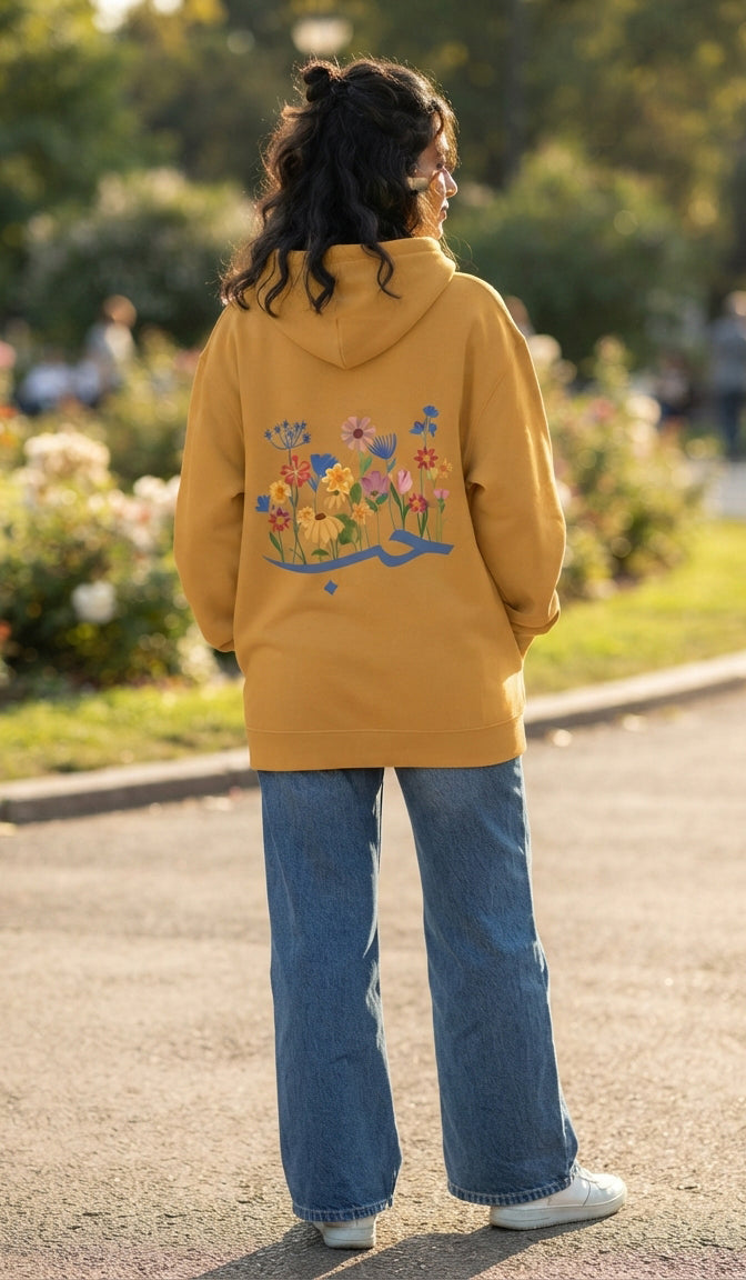 A person standing outdoors on a paved path, viewed from behind, wearing a mustard yellow hoodie with a colorful floral and bird graphic on the back, blue jeans, and white sneakers.