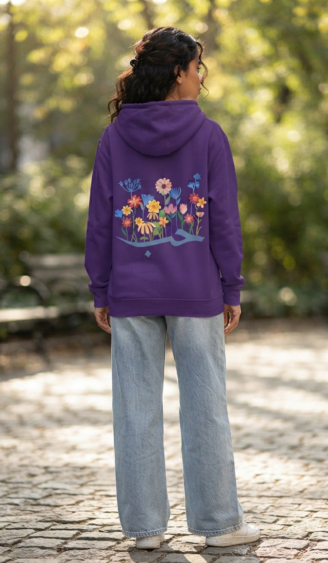 A woman viewed from behind wearing a purple hoodie with a colorful floral graphic on the back, paired with light blue jeans, standing on a cobblestone path in a sunlit park.