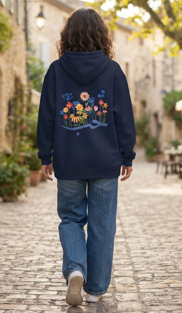 A person viewed from behind walking down a cobblestone street, wearing a navy blue hoodie with a colorful floral and wave graphic on the back, paired with blue jeans.