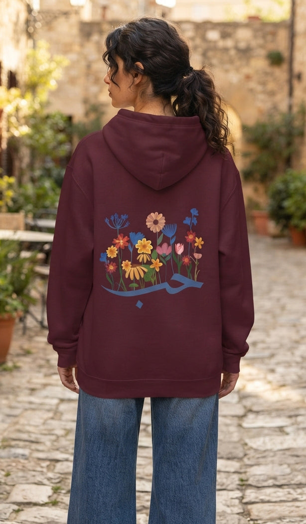 A woman viewed from behind wearing a burgundy hoodie with a colorful embroidered floral design on the back, paired with blue jeans, standing on a stone pathway.