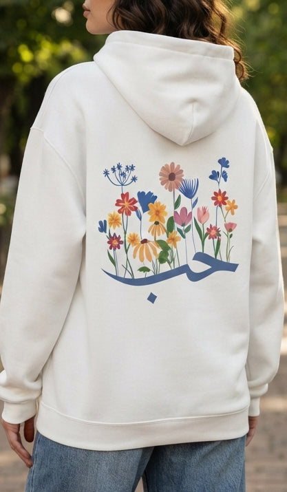 A person wearing an off-white hoodie from the back, featuring a colorful floral and Arabic calligraphy graphic on the back, paired with blue jeans in an outdoor setting.