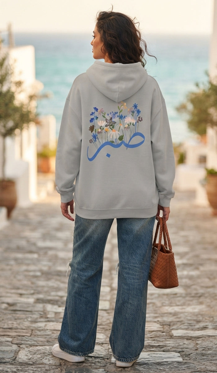A woman viewed from behind wearing a light gray hoodie with a large blue Arabic calligraphy graphic and a floral print on the back, paired with wide-leg blue jeans and holding a brown woven handbag on a cobblestone street with the sea in the background.