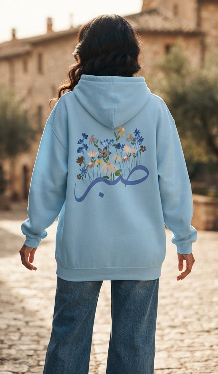 A person viewed from behind wearing a light blue hoodie with a large floral graphic and Arabic calligraphy on the back, paired with blue jeans, standing outdoors on a cobblestone street.