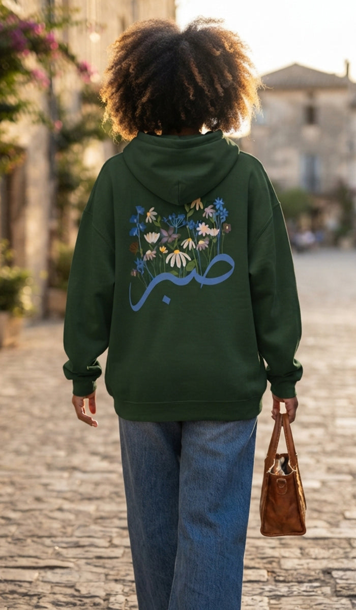A person viewed from behind wearing a dark green hoodie with a colorful floral and blue Arabic calligraphy graphic on the back, paired with blue jeans and holding a brown handbag on a cobblestone street.