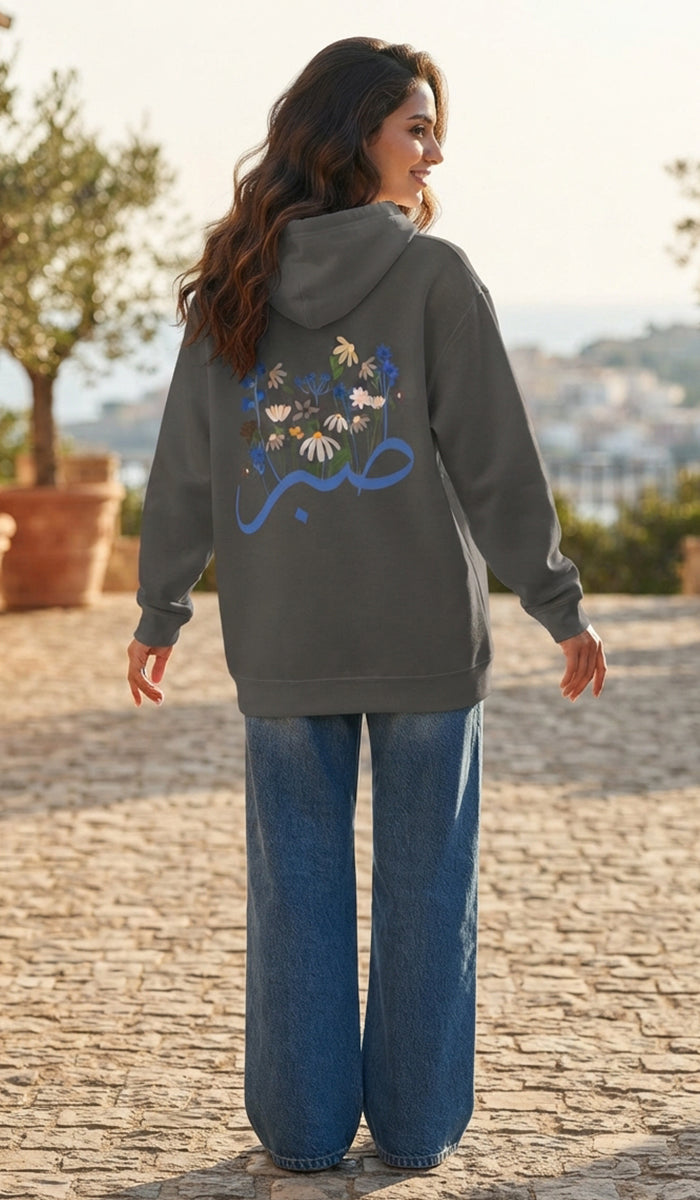 A woman stands outdoors on a cobblestone path wearing a dark gray hoodie with a colorful floral and script graphic on the back, paired with wide-leg blue jeans.