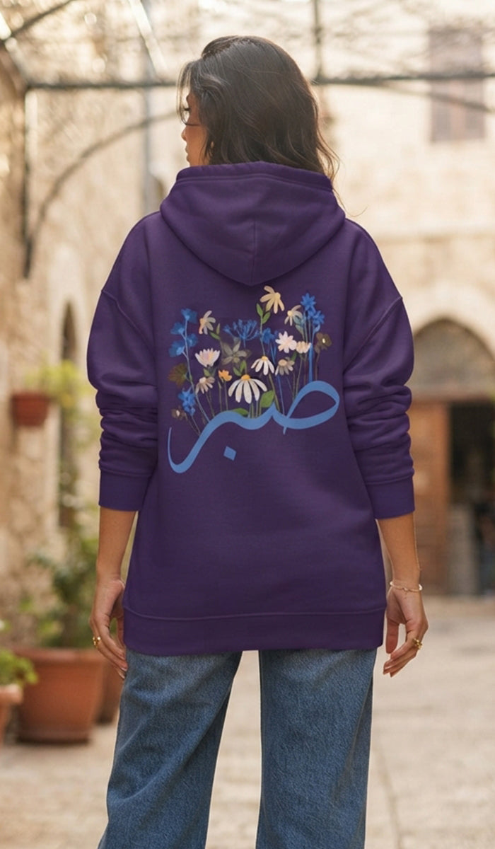 A woman viewed from behind wearing a purple hoodie with a large blue and multicolor floral graphic and Arabic calligraphy on the back, paired with blue jeans in an outdoor courtyard setting.