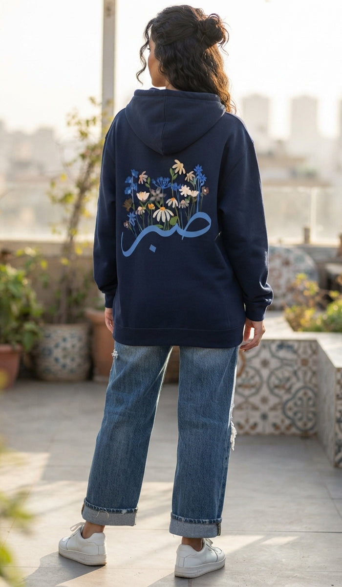 A woman viewed from behind wearing a navy blue hoodie with a large floral graphic and Arabic calligraphy on the back, paired with wide-leg blue jeans and white sneakers, standing on an outdoor rooftop.