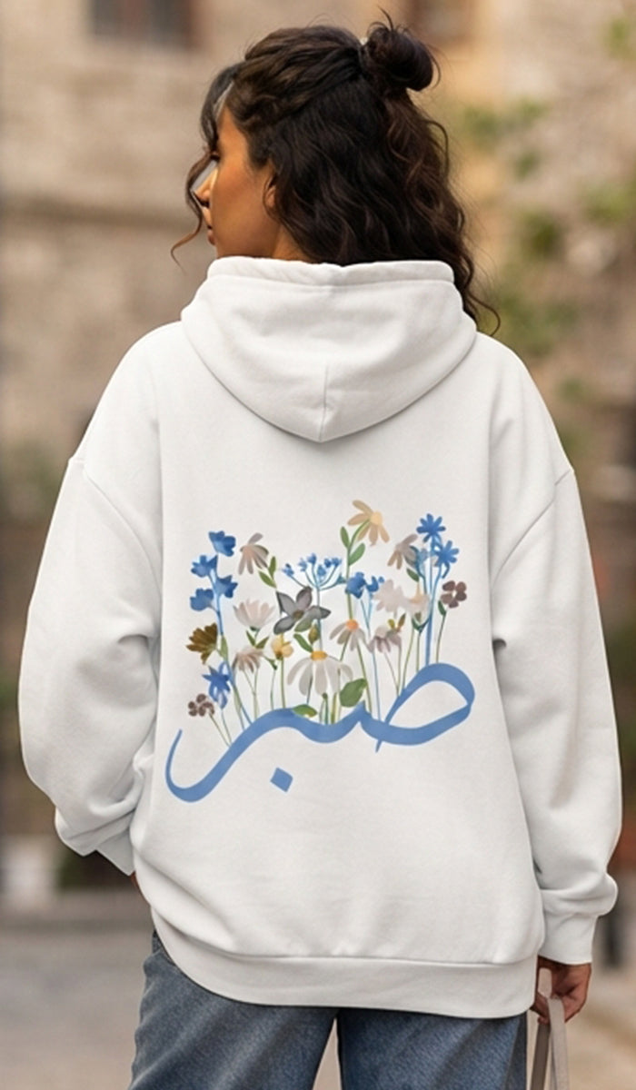 A woman viewed from behind wearing a white hoodie with a colorful floral and Arabic calligraphy graphic on the back, paired with blue jeans in an outdoor setting.