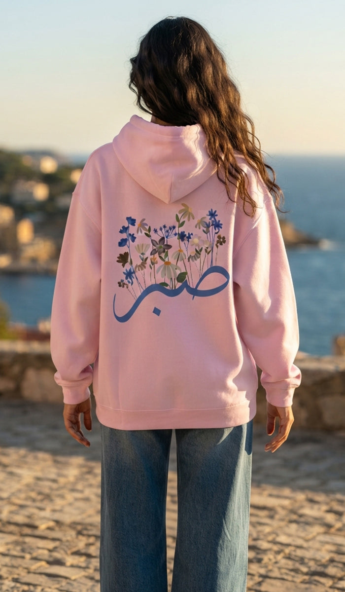 A woman viewed from behind wearing a light pink hoodie with a large blue Arabic calligraphy graphic and a floral design on the back, paired with wide-leg blue jeans, set against a coastal background.