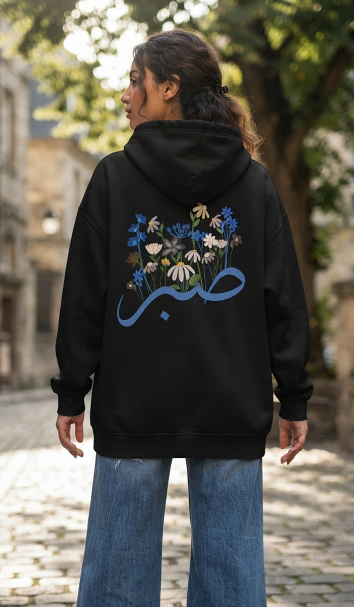 A woman wearing a black hoodie with a large blue Arabic calligraphy graphic and a floral design on the back, paired with wide-leg blue jeans, standing on a cobblestone street.