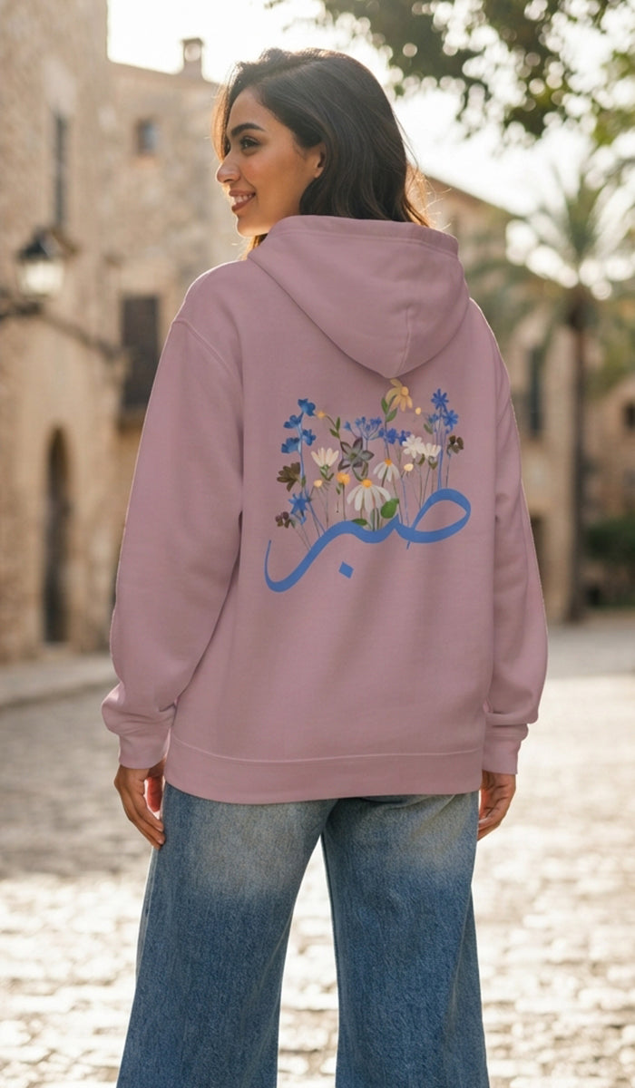 A woman wearing a dusty pink hoodie with a blue Arabic calligraphy graphic and a floral design on the back, paired with blue jeans, standing in an outdoor street setting.