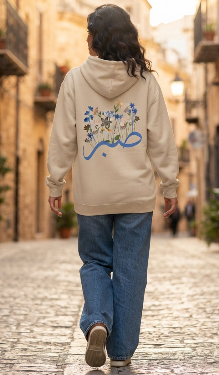 A woman viewed from behind walking down a cobblestone street, wearing a beige hoodie with a blue Arabic calligraphy graphic and floral print on the back, paired with blue jeans.
