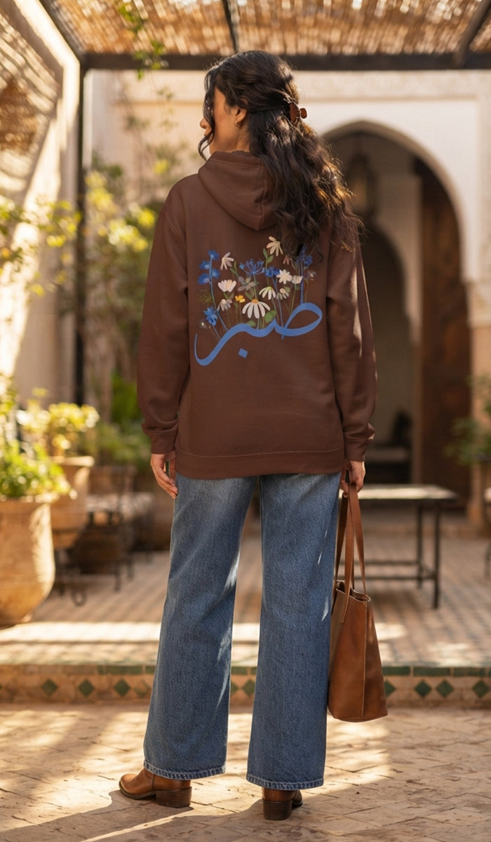 A woman stands in a sunlit courtyard wearing a brown hoodie with a large multicolor floral and script graphic on the back, paired with wide-leg blue jeans and brown boots. She is holding a brown tote bag.