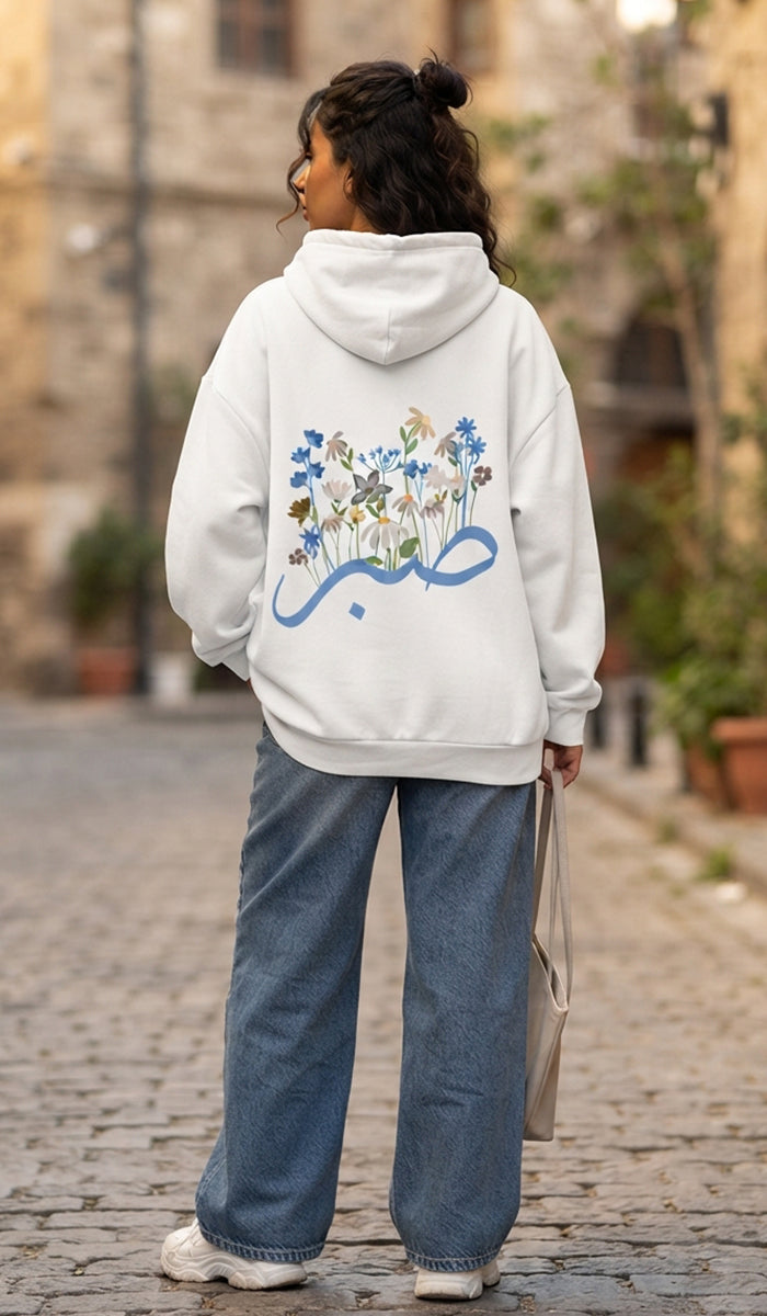 A woman viewed from behind wearing a white hoodie with a blue Arabic calligraphy graphic and a floral design on the back, paired with wide-leg blue jeans and white sneakers, standing on a cobblestone street.