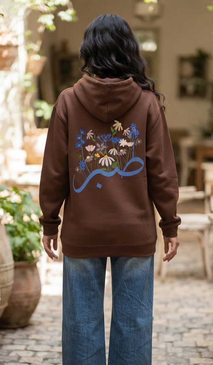 A woman viewed from behind wearing a brown hoodie with a colorful floral and blue Arabic calligraphy graphic on the back, paired with blue jeans in an outdoor patio setting.