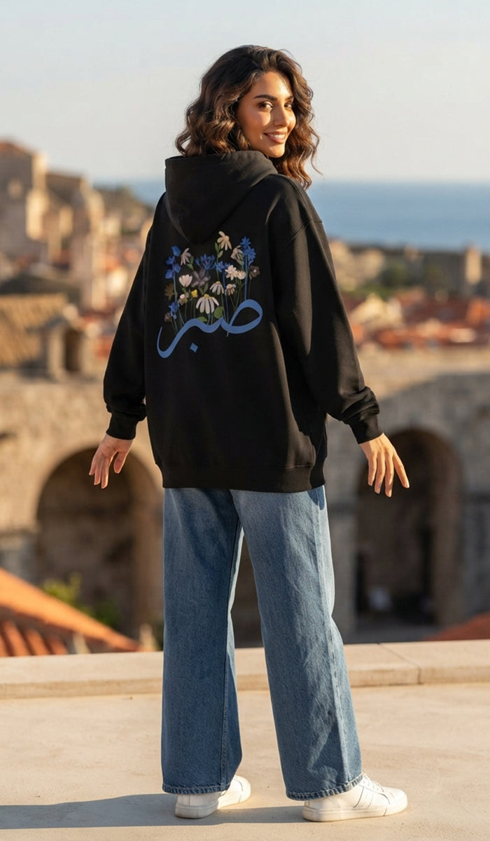 A woman wearing a black hoodie with a blue Arabic calligraphy graphic and floral print on the back, paired with wide-leg blue jeans and white sneakers, standing outdoors with a blurred cityscape background.