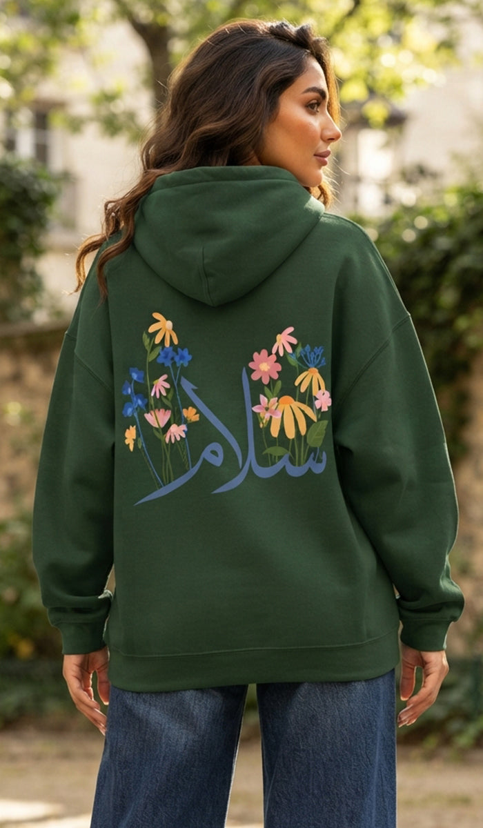 A woman wearing an olive green hoodie with colorful floral embroidery and Arabic calligraphy on the back, paired with blue jeans, photographed outdoors with a blurred background.