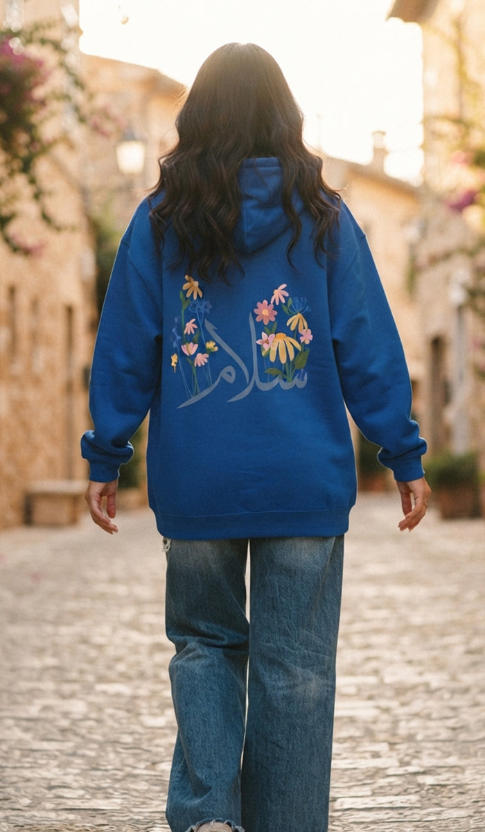 A woman viewed from behind wearing a royal blue hoodie with colorful floral embroidery and Arabic calligraphy on the back, paired with wide-leg blue jeans, walking on a sunlit cobblestone street.