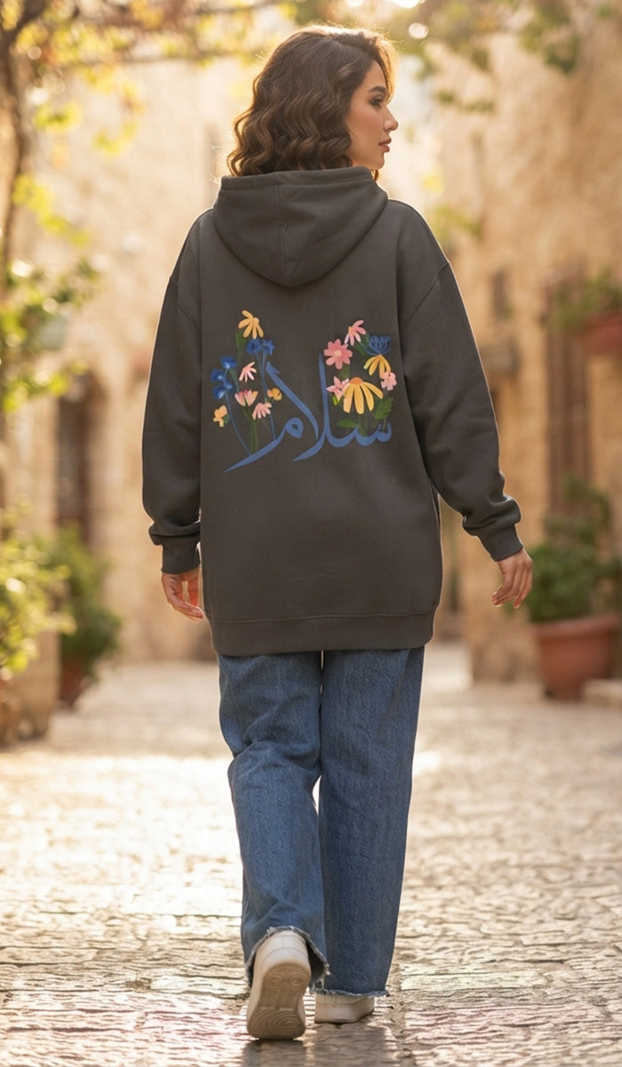 A woman walking down a sunlit cobblestone street, viewed from behind. She is wearing a dark charcoal gray hoodie with colorful floral embroidery and blue jeans.