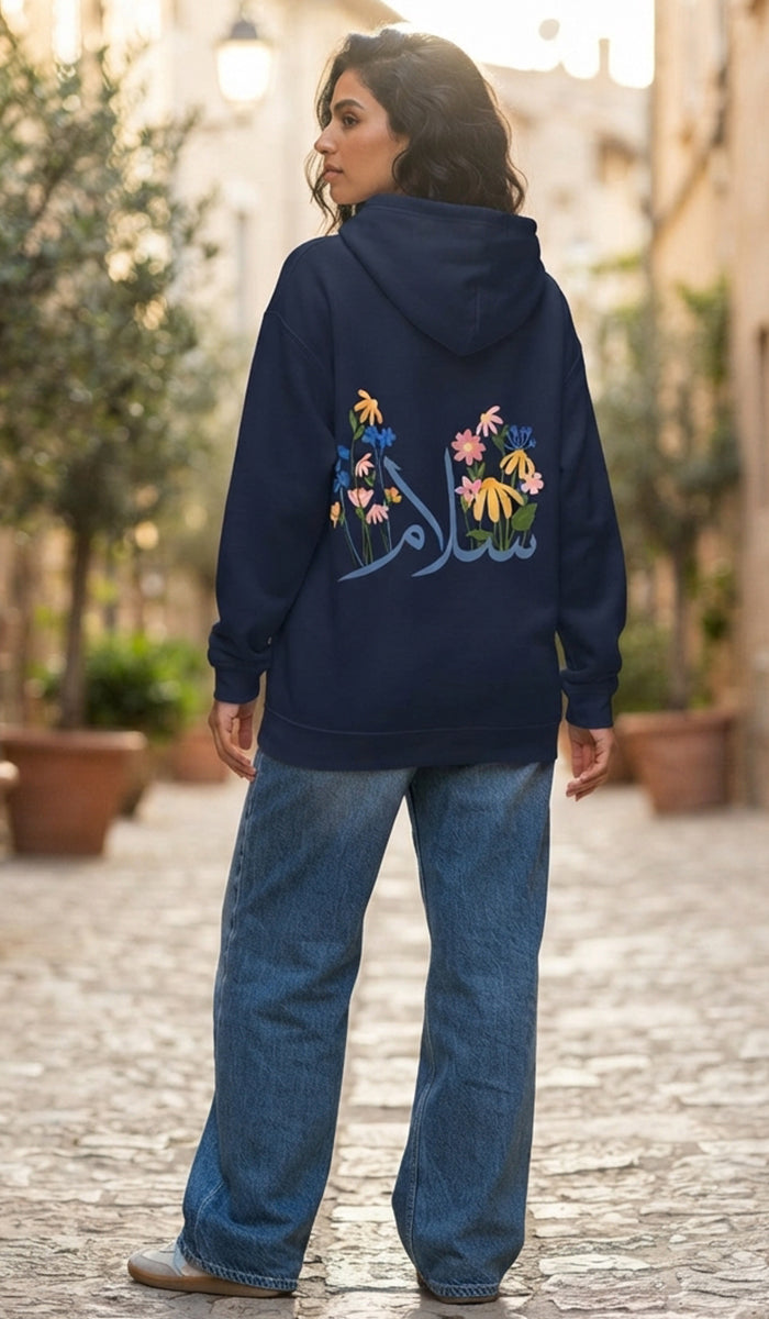 A woman wearing a navy blue hoodie with colorful floral embroidery and Arabic calligraphy on the back, paired with wide-leg blue jeans, standing outdoors on a cobblestone path.