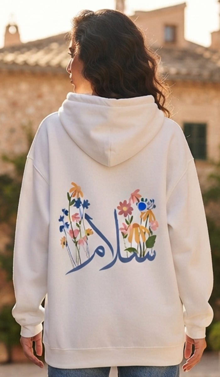 A woman viewed from behind wearing an oversized white hoodie with colorful floral graphics and Arabic calligraphy on the back, set against a warm, blurred outdoor background of stone buildings.