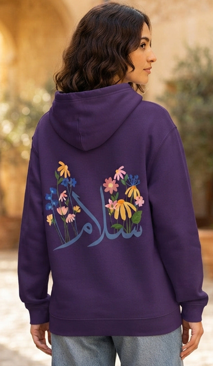 A woman wearing a purple hoodie with colorful floral embroidery and Arabic calligraphy on the back, shown from behind in an outdoor setting.