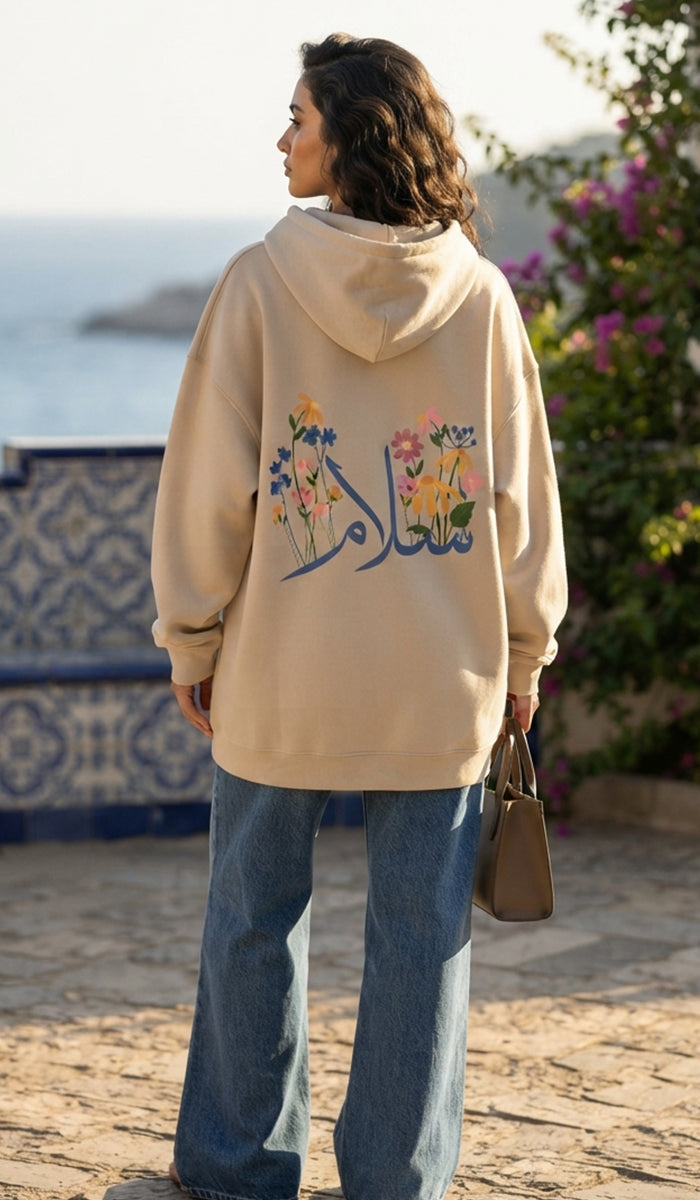A woman stands outdoors by the sea wearing a beige oversized hoodie with colorful floral embroidery and Arabic calligraphy on the back, paired with wide-leg blue jeans and holding a brown handbag.