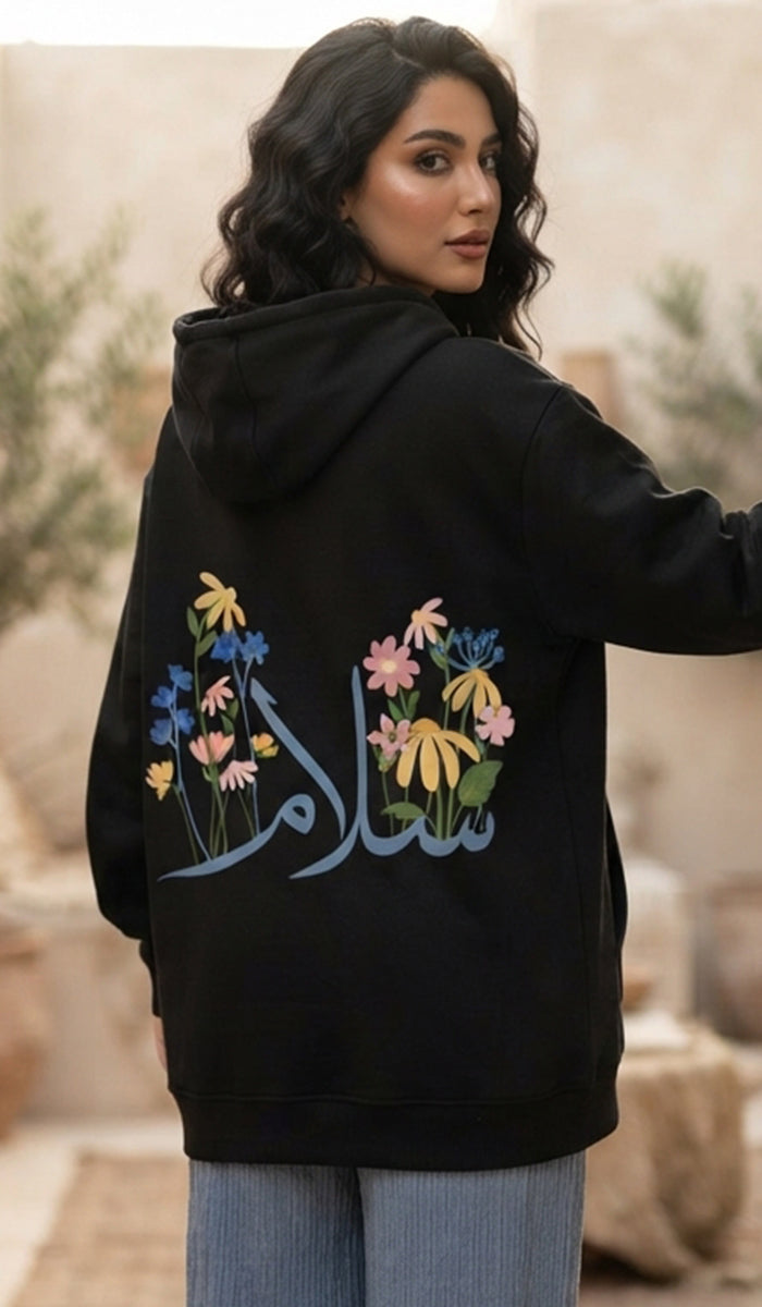 A woman wearing a black hoodie with colorful floral embroidery and Arabic calligraphy on the back, paired with light blue jeans, photographed in a warm, neutral-toned outdoor setting.