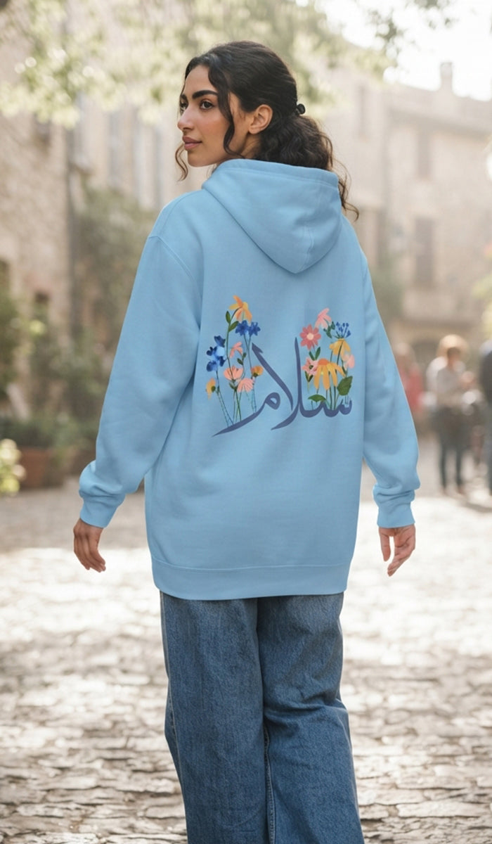 A woman wearing a light blue hoodie with colorful floral embroidery and Arabic calligraphy on the back, paired with wide-leg blue jeans, standing on a cobblestone street.
