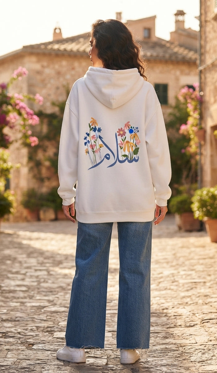 A woman viewed from behind wearing a white hoodie with colorful floral embroidery and Arabic calligraphy on the back, paired with wide-leg blue jeans, standing on a cobblestone street in front of a sunlit stone building.