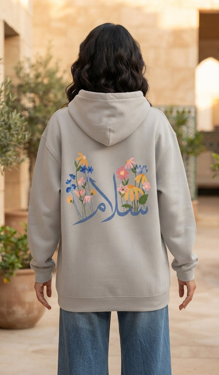 A person viewed from behind wearing a light beige hoodie with a colorful floral and Arabic calligraphy graphic on the back, paired with blue jeans in an outdoor courtyard setting.
