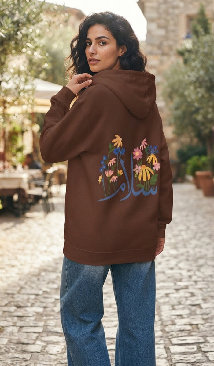 A woman wearing a brown hoodie with colorful floral embroidery and Arabic calligraphy on the back, paired with blue jeans, standing on a cobblestone street.