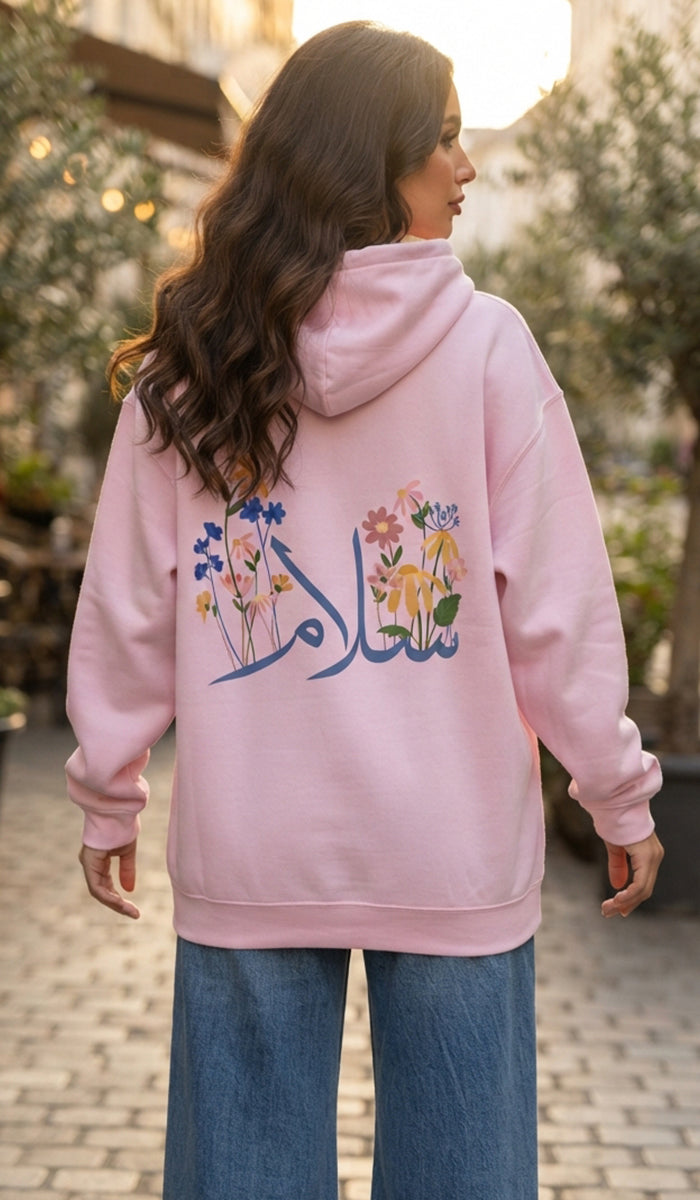 A woman viewed from behind wearing a light pink hoodie with a colorful floral graphic and Arabic calligraphy on the back, paired with blue jeans in an outdoor setting.