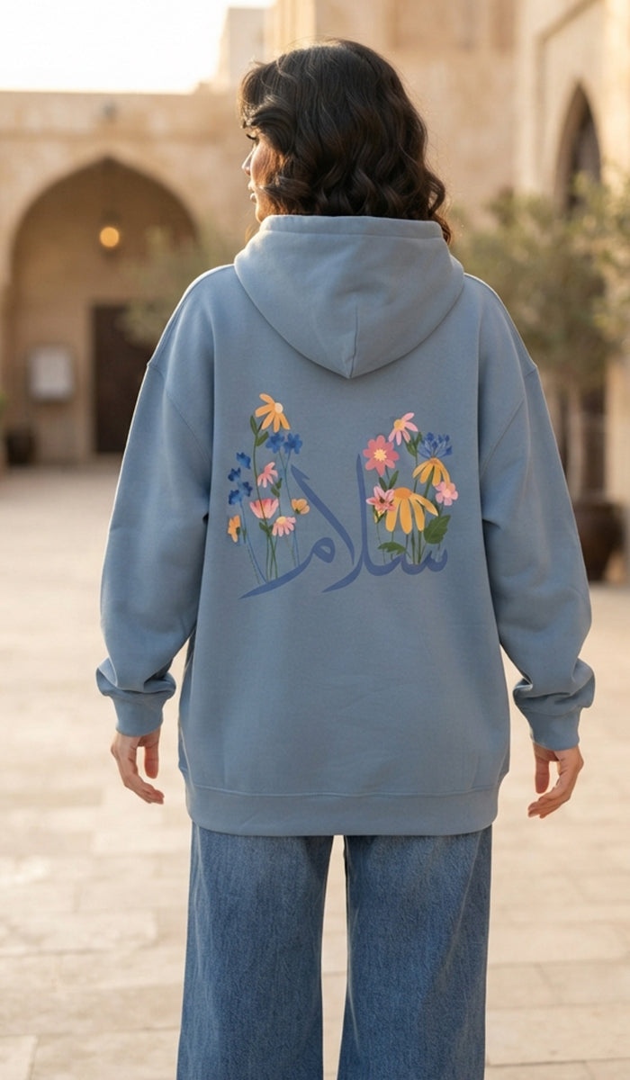 A woman viewed from behind wearing a dusty blue hoodie with colorful floral embroidery and Arabic calligraphy on the back, paired with wide-leg blue jeans in an outdoor courtyard setting.