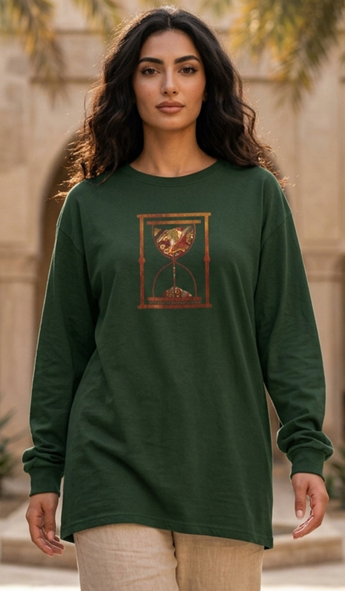 A woman wearing a dark forest green long-sleeve tunic top with a gold and brown hourglass graphic on the chest, paired with beige pants, standing outdoors against a warm stone background.