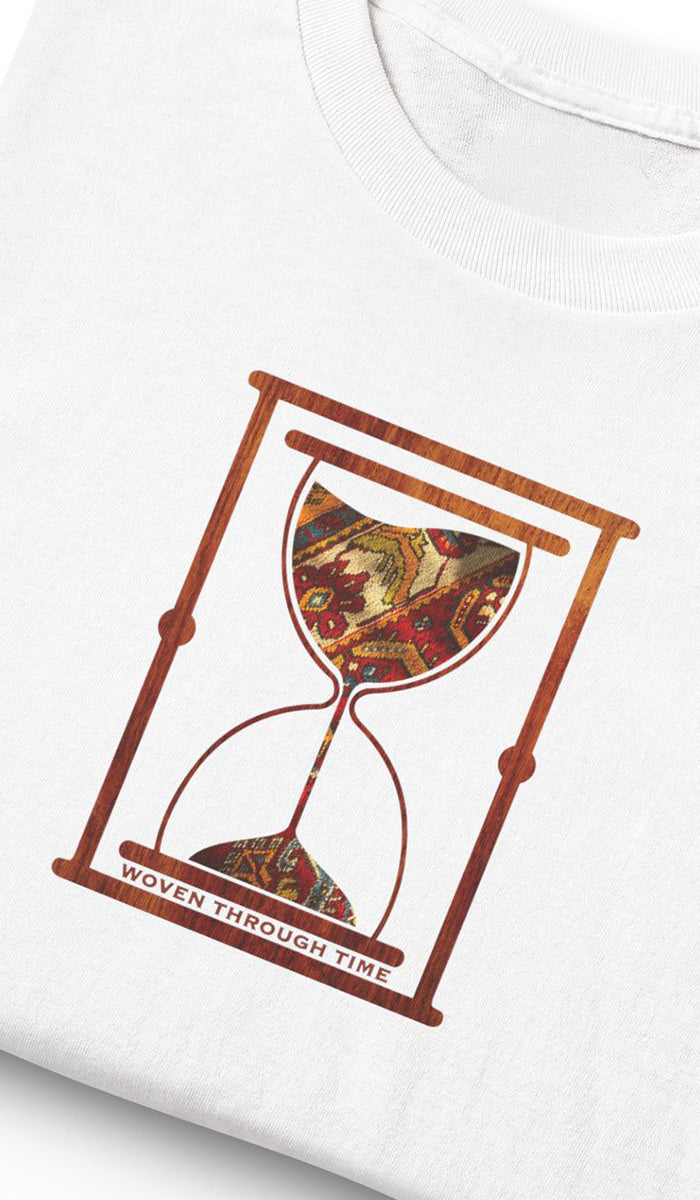 Close-up of a white T-shirt with a graphic print of an hourglass inside a rectangular frame, featuring a detailed woven pattern in red, gold, and brown, with the text 'WOVEN THROUGH TIME' along the bottom.