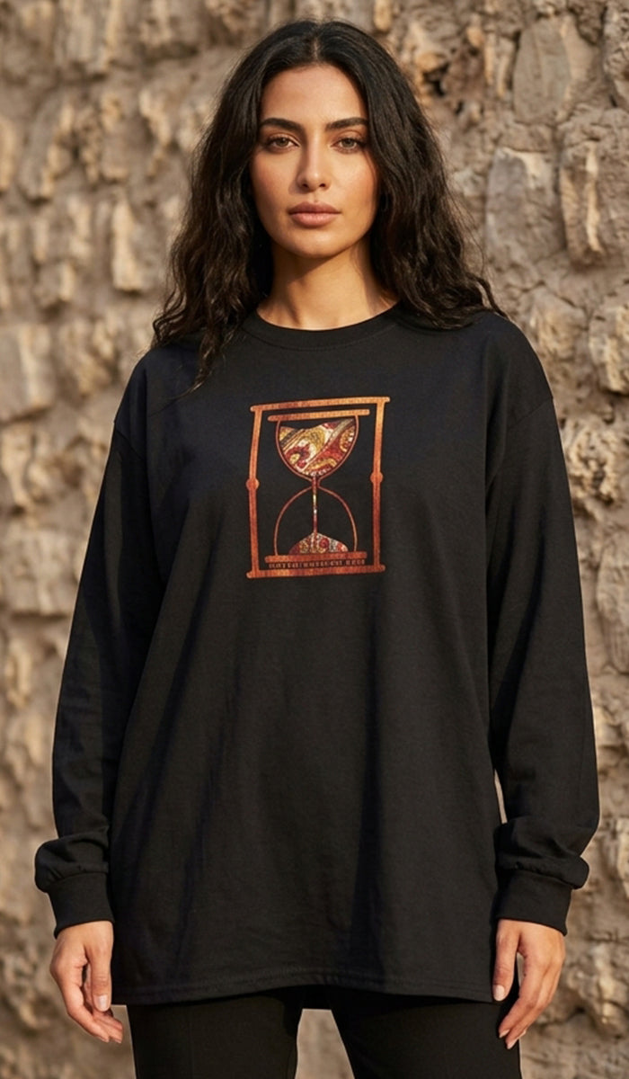A woman wearing a black long-sleeve t-shirt with a red and gold hourglass graphic on the chest, standing against a rustic stone wall.