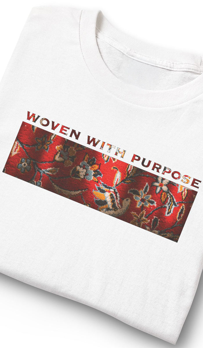 A folded white crewneck t-shirt featuring a graphic print with the text 'WOVEN WITH PURPOSE' in red and a red patterned rectangular panel with traditional Asian-style motifs.