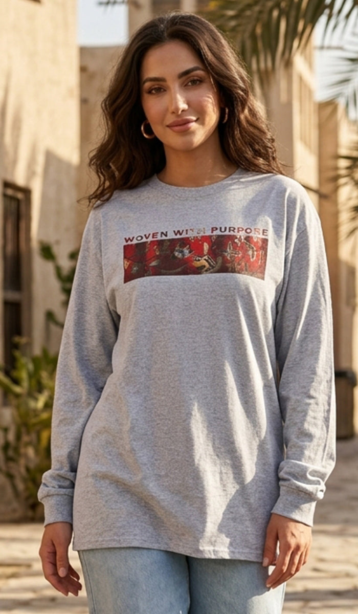 A woman wearing a light heather gray long-sleeve t-shirt with a red graphic print on the chest that reads 'WOVEN WITH PURPOSE'. She is also wearing light blue jeans, standing outdoors in a sunny, warm-toned environment.