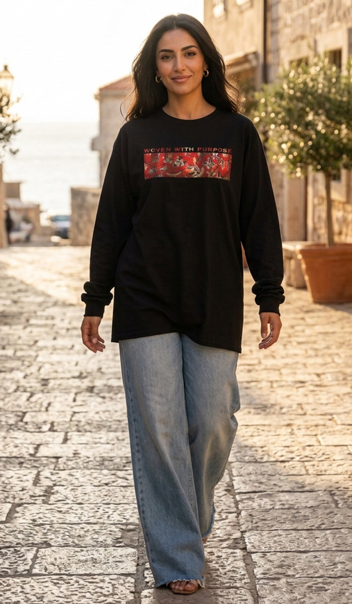A woman walking on a cobblestone street wearing a black long-sleeve graphic sweatshirt with a red and gold chest print, paired with light-wash wide-leg jeans.