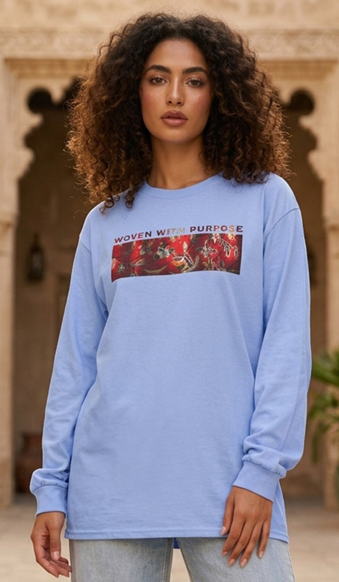 A woman with curly brown hair wearing a light blue long-sleeve graphic sweatshirt with the text 'WOVEN WITH PURPOSE' and a red graphic print on the chest, standing in front of a stone archway.