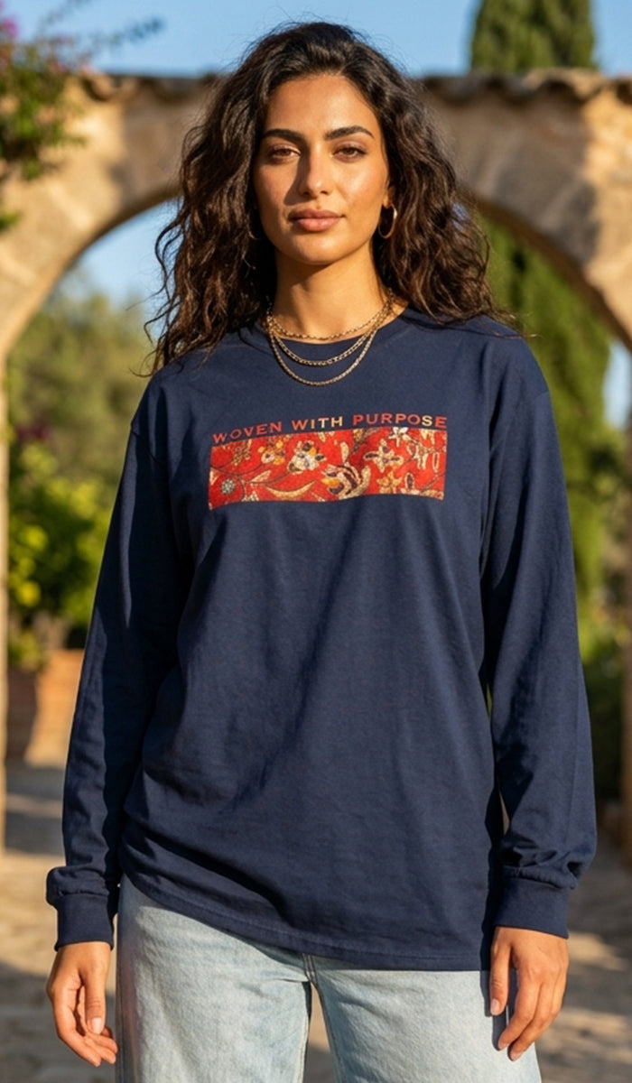 A woman wearing a navy blue long-sleeve t-shirt with a red graphic print on the chest that reads 'WOVEN WITH PURPOSE'. She is standing outdoors in front of a stone archway.