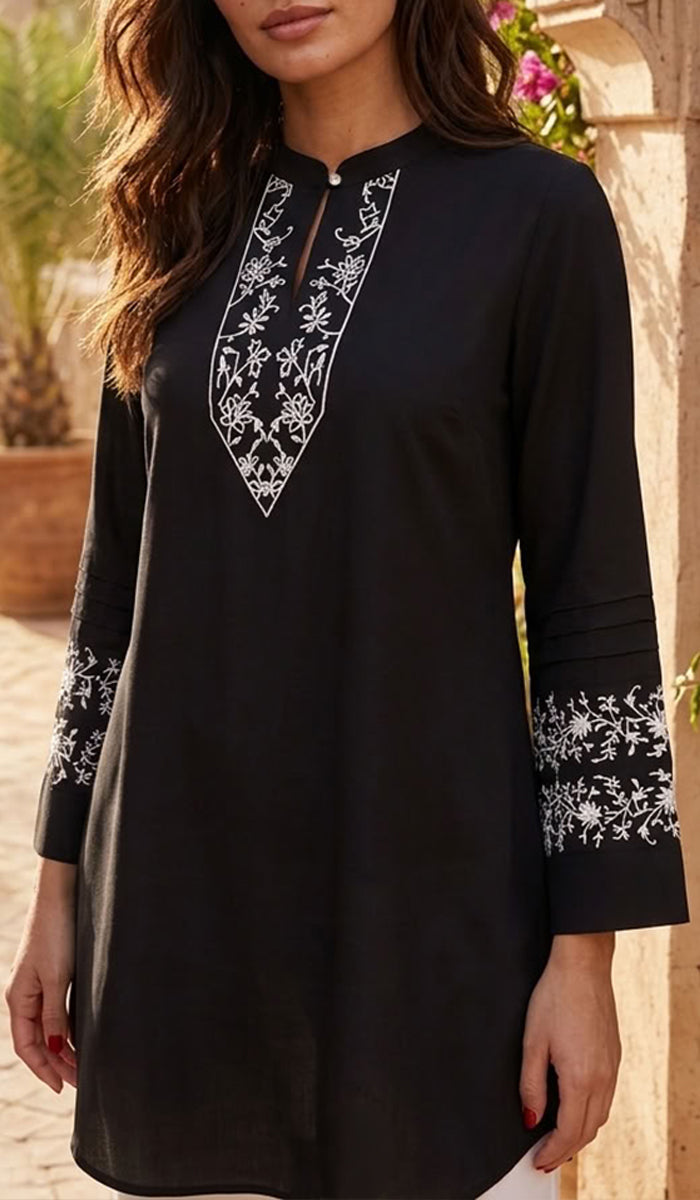 A woman wearing a black long-sleeve tunic with white floral embroidery around the V-neckline and cuffs, photographed outdoors against a warm stone wall background.