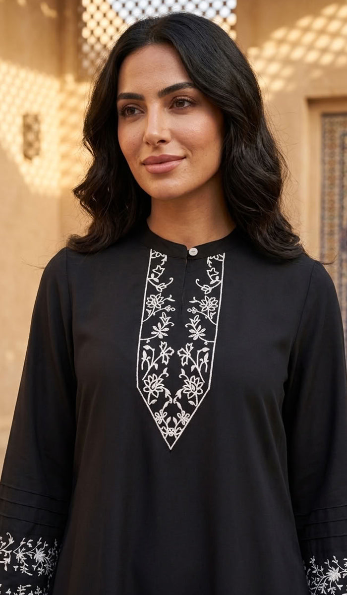 A woman wearing a black long-sleeve tunic with white floral embroidery around the V-neckline and sleeve cuffs, set against a warm-toned architectural background.