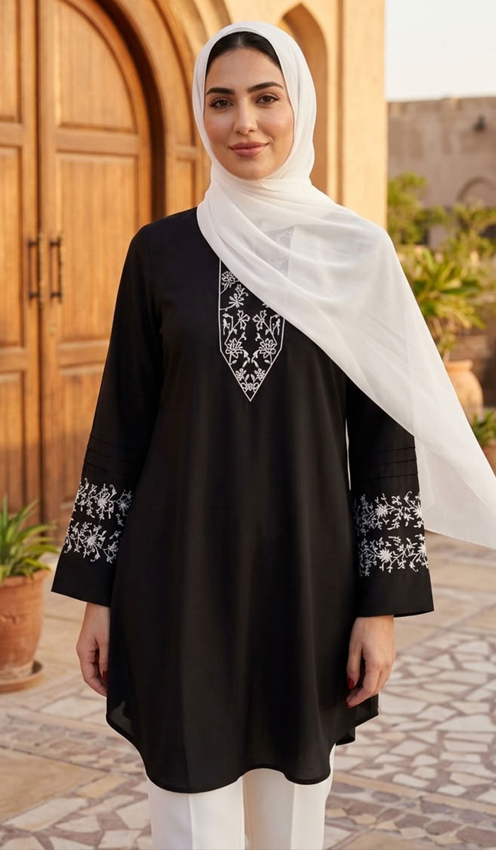A woman wearing a black long-sleeve tunic with white floral embroidery on the neckline and cuffs, paired with a white hijab and white pants, standing in front of a warm-toned wooden door.