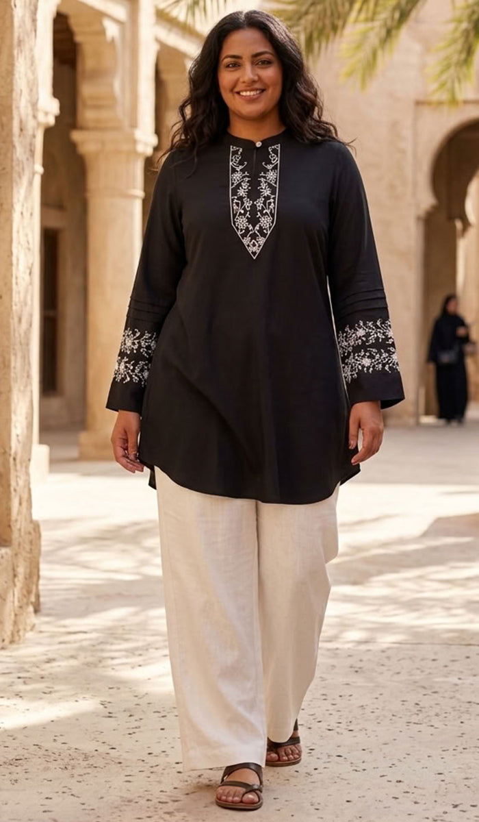 A woman walking outdoors in a black kurta with white embroidery on the neckline and cuffs, paired with white wide-leg pants and brown sandals.