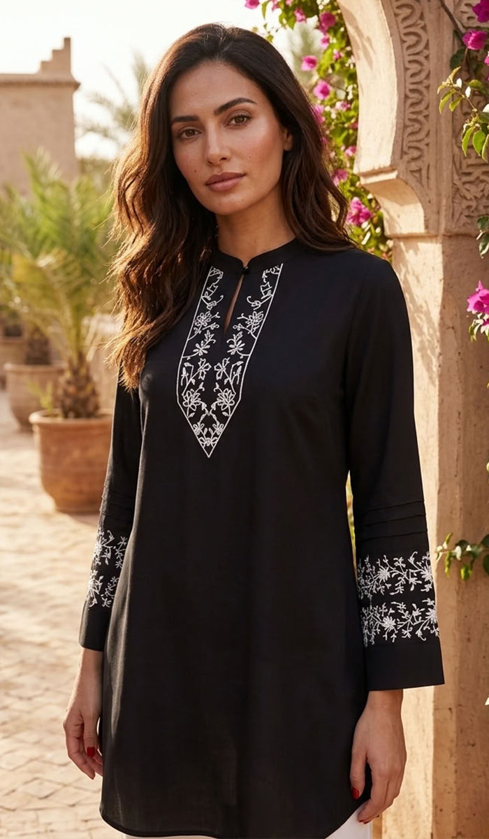 A woman wearing a black tunic with white floral embroidery on the neckline and cuffs, standing outdoors in front of a stone archway with plants and flowers.
