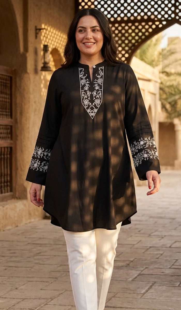 A woman wearing a black long kurta with white floral embroidery on the neckline and cuffs, paired with white pants, walking outdoors in front of a traditional stone archway.
