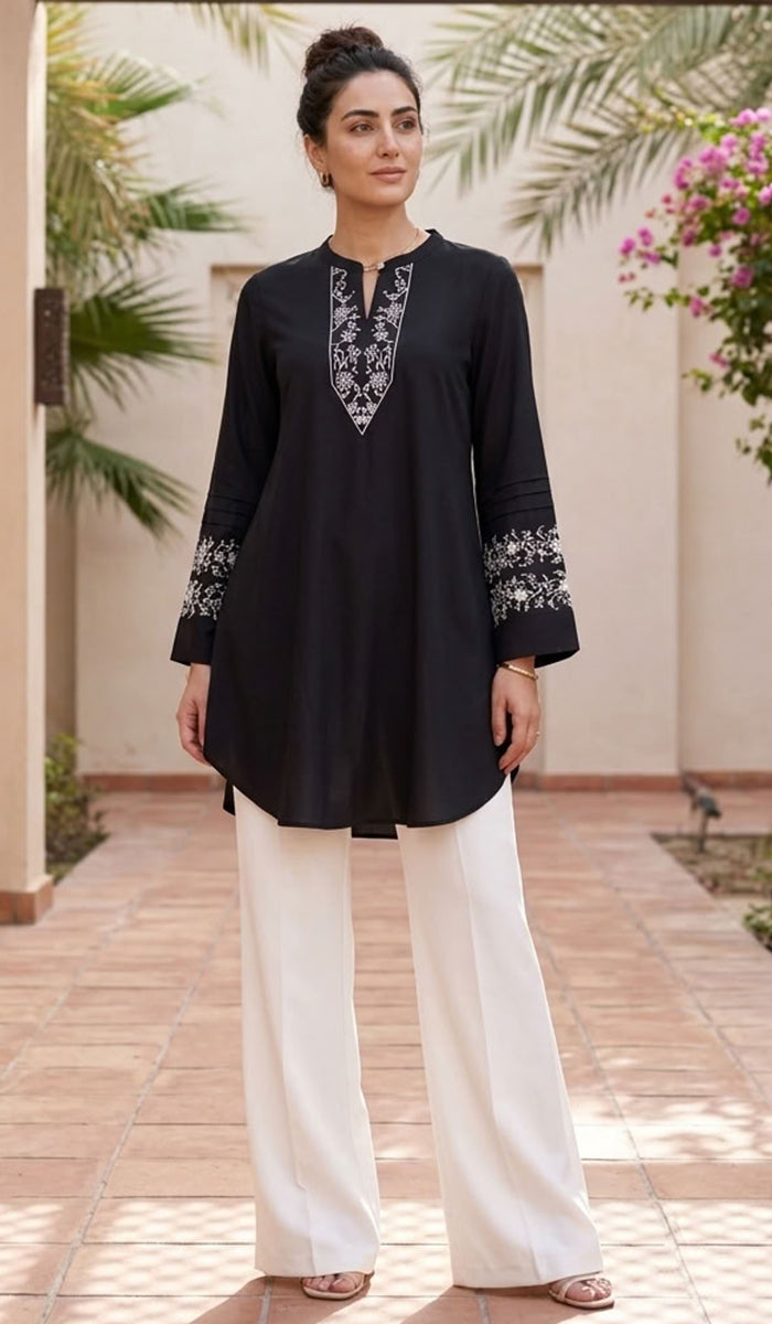 A woman modeling a black long-sleeve kurta with intricate silver and white embroidery on the neckline and cuffs, paired with white wide-leg trousers, set against a warm outdoor courtyard background with palm trees.