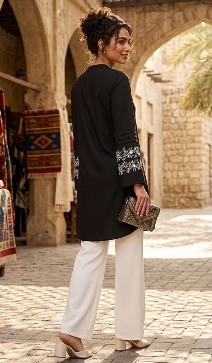 A woman stands in a sunlit outdoor market walkway wearing a black long kurta with white embroidered cuffs, white wide-leg pants, and beige heels, while holding a small clutch.